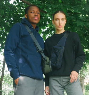 A male and female model stand in front of a leafy green tree, each wearing a sweater and a bag from Bedi
