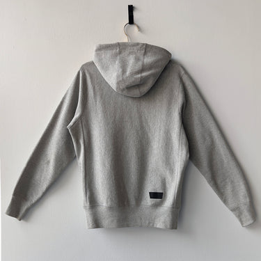 Gray hoodie with a small black logo on the back, hanging against a white wall.