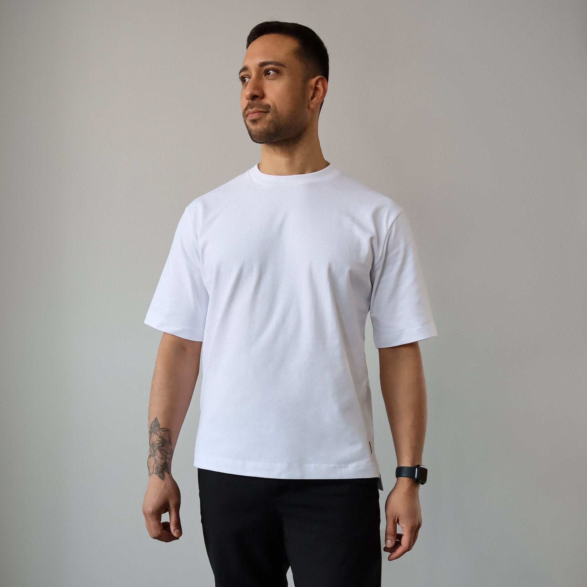 A man stands casually in front of a plain wall, wearing the white Blake tshirt and black pants