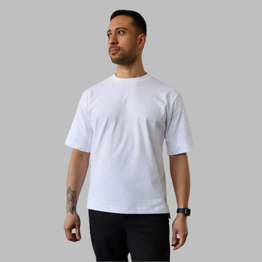 A man stands casually in front of a plain wall, wearing the white Blake tshirt and black pants