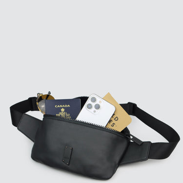 A black nylon fanny pack with a phone, passport, notebook and sunglasses against a white background