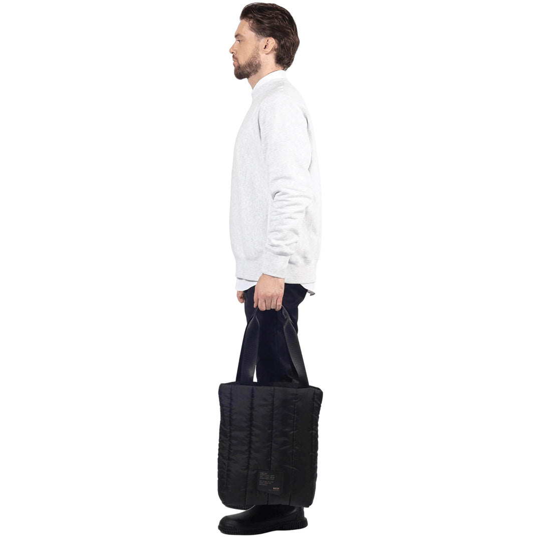 Male model stands, holding a utilitarian style medium sized tote back in black quilted deadstock material, on a white background.