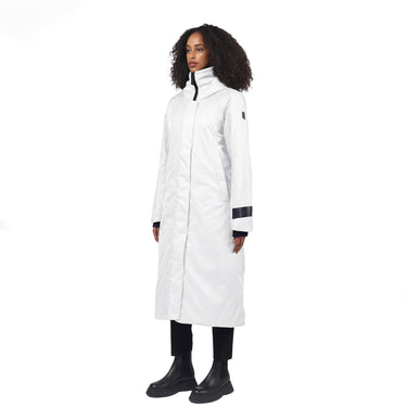 A woman modeling a long white coat with black trim details on the cuffs and collar, paired with black pants and boots, against a plain white background.