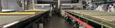 A view down the aisle between two cutting tables in a small local garment workshop. A man works in the distance at the end of the aisle