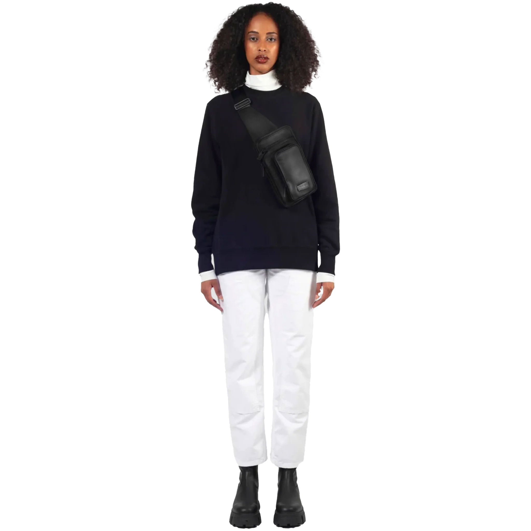 Female model stands, wearing the sling sling in black upcycled leather with a sleek finish as a crossbody on a white background.