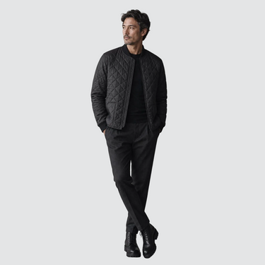 A man modeling a black quilted bomber jacket over a black turtleneck, black trousers, and black leather boots, standing against a solid light gray background.