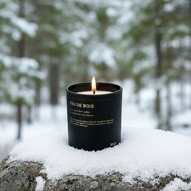 Black candle labeled 'Feu de Bois' on a snowy stone surface with a forest background