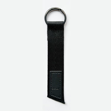 A simple all-black keychain made from a black seatbelt, with black metal ring and a faux leather logo patch with black letters