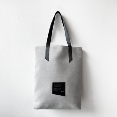 A white tote bag made with thick, textured material and a black strap hangs against a white wall