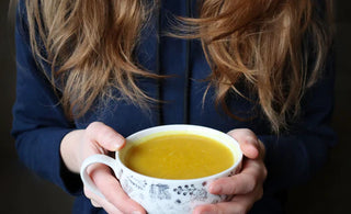 A woman with long red hair wears a navy Bedi hoodie, and holds a mug of turmeric latte in her hands
