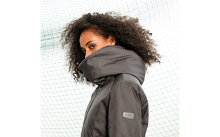 A female model burries the bottom part of her face in the high neck/hood of an evergreen coat, and looks sideways at the camera