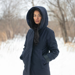 A woman stands in a snowy field, wearing a navy winter coat with the hood up.