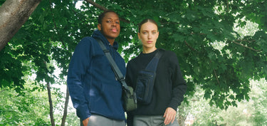 A male and female model stand in front of a leafy green tree, each wearing a sweater and a bag from Bedi