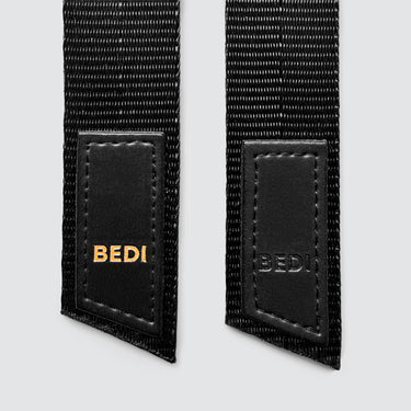a close up of the bottom of two keychains made with seatbelts, both with the bedi logo, one with gold lettering and one with black lettering. 