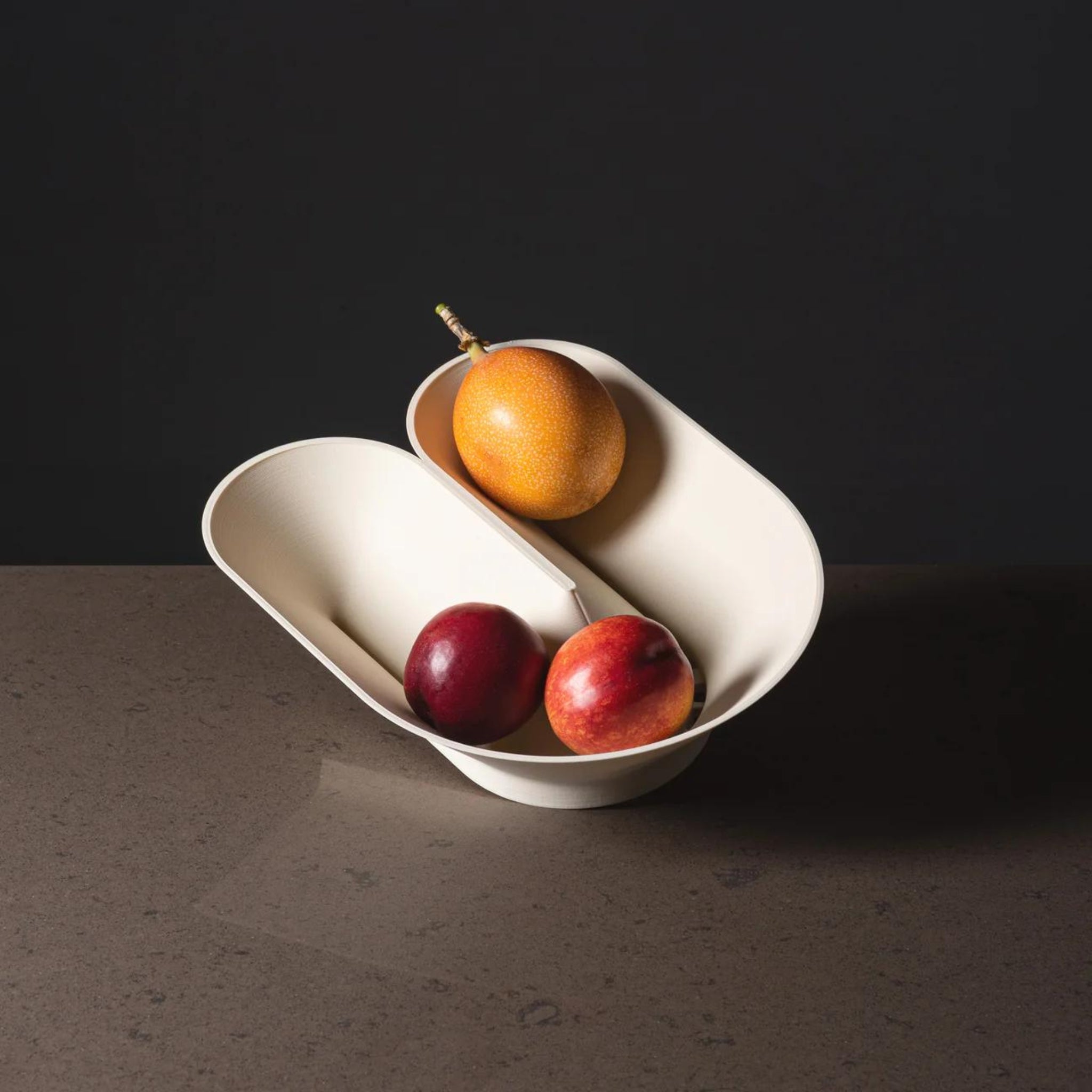 Cyrc U fruit bowl in eggshell. 3D printed recycled plastic fruit bowl holding three red fruits, resting on a granite table in front of a dark backgound.
