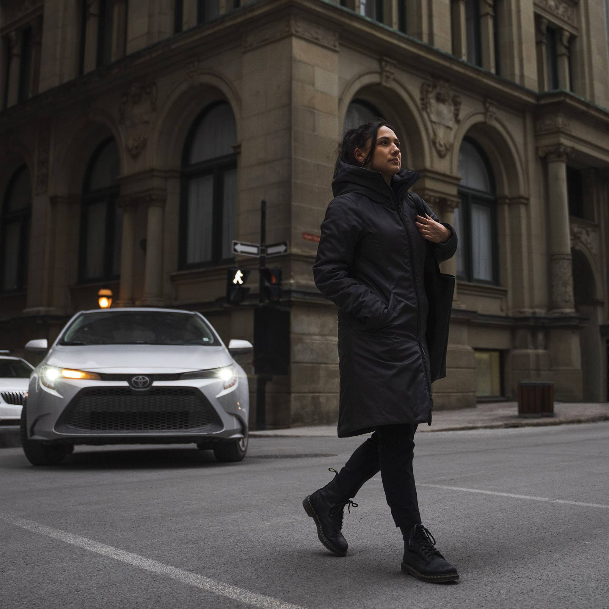 A woman confidently crosses the street wearing the Kirsi, a knee-length coat, in black
