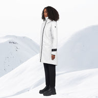 A woman stands in a snowy mountain landscape wearing a long white parka with black trim and black pants.