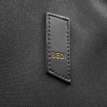 Close-up of a black textured fabric surface with a stitched black apple leather patch embossed with the gold-tone BEDI brand name.