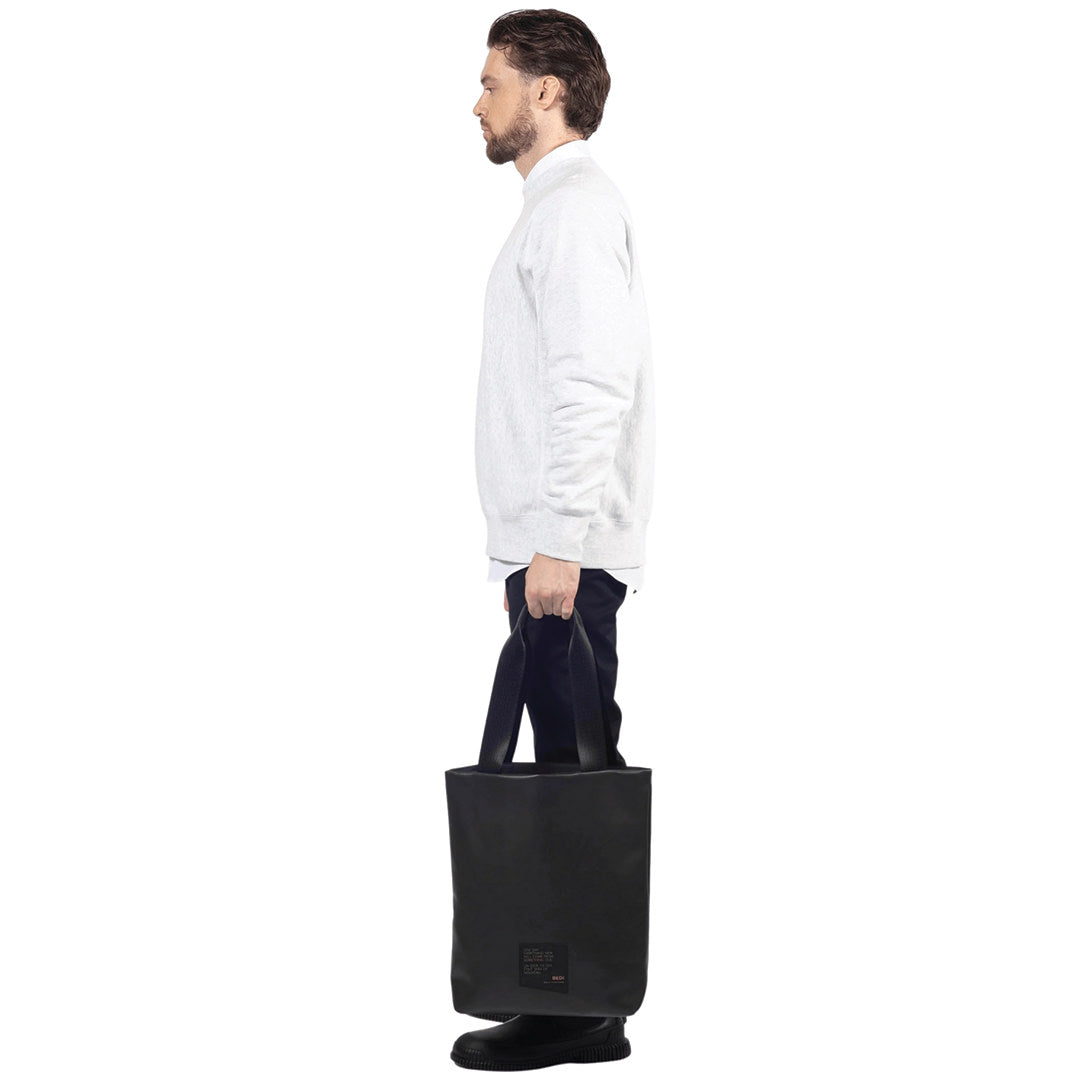 Male model stands, holding on his side a utilitarian style medium sized tot bag in vegan leather (Desserto), on white background.