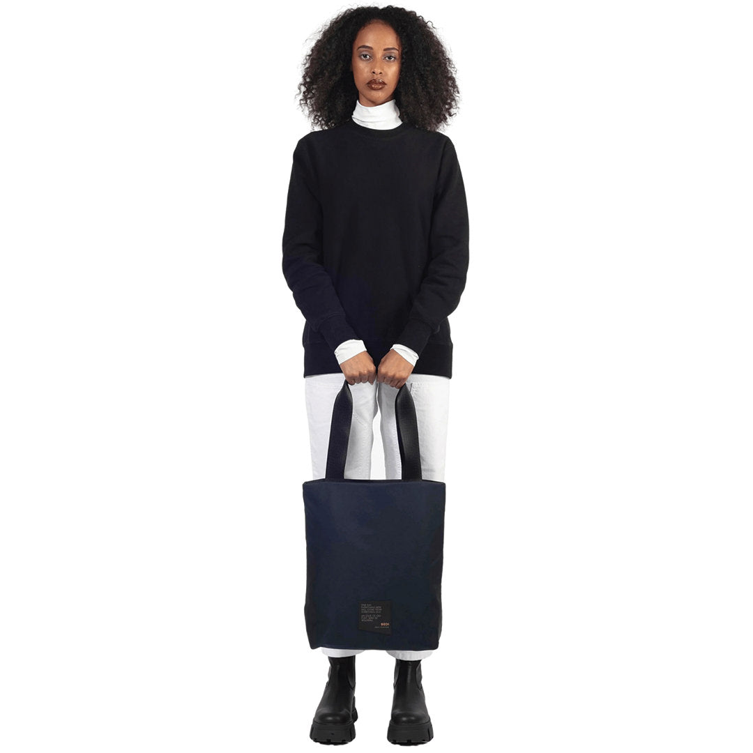 Female model stands, holding a utilitarian style medium sized tote bag in navy deadstock nylon, on white background.