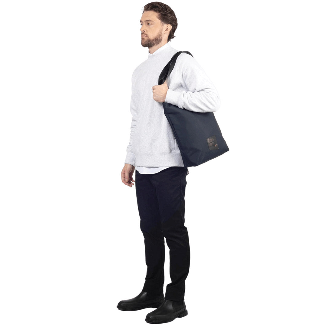 Male model stands, holding on his left side shoulder a utilitarian style medium sized tote bag in navy deadstock nylon, on white background.