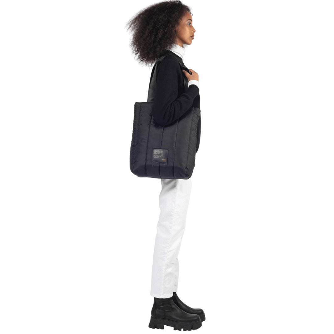 Female model stands, holding a utilitarian style medium sized tote in black quilted deadstock material, on a white background.