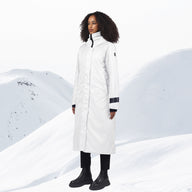 A woman stands in a snowy mountain landscape wearing a long white parka with black trim at the cuffs and collar, paired with black pants and boots.