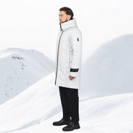 A man wearing a long white insulated parka with black trim, black pants, and black boots, standing in a snowy mountain landscape.