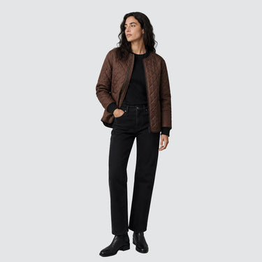 A woman modeling a brown quilted bomber jacket over a black top and black straight-leg jeans, standing against a solid light gray studio background.