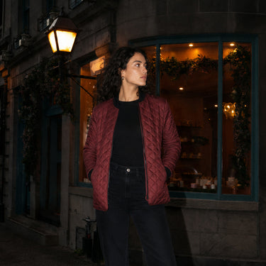 A woman stands outdoors at night in front of a storefront window, wearing a quilted burgundy jacket over a black top and dark jeans. A lit street lamp and the shop interior are visible in the background.