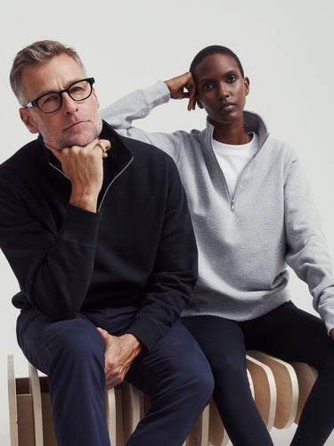 A man and a woman both wearing 1/4 zip sweaters, sitting on a wooden bench with a plain background