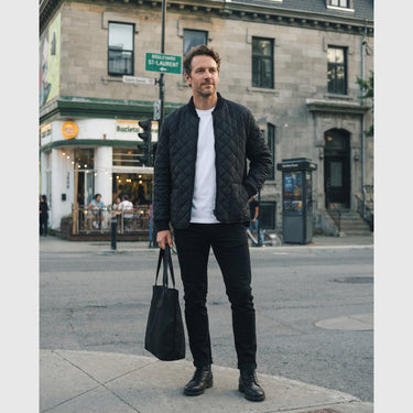 A man stands on a city street wearing a black quilted bomber jacket over a white T-shirt, black pants, and black shoes, while holding a black tote bag. Buildings and a street sign are visible in the background.
