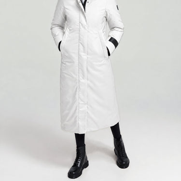 A model wearing a long white winter coat with black trim details, paired with black socks and black boots, photographed against a light grey studio background.