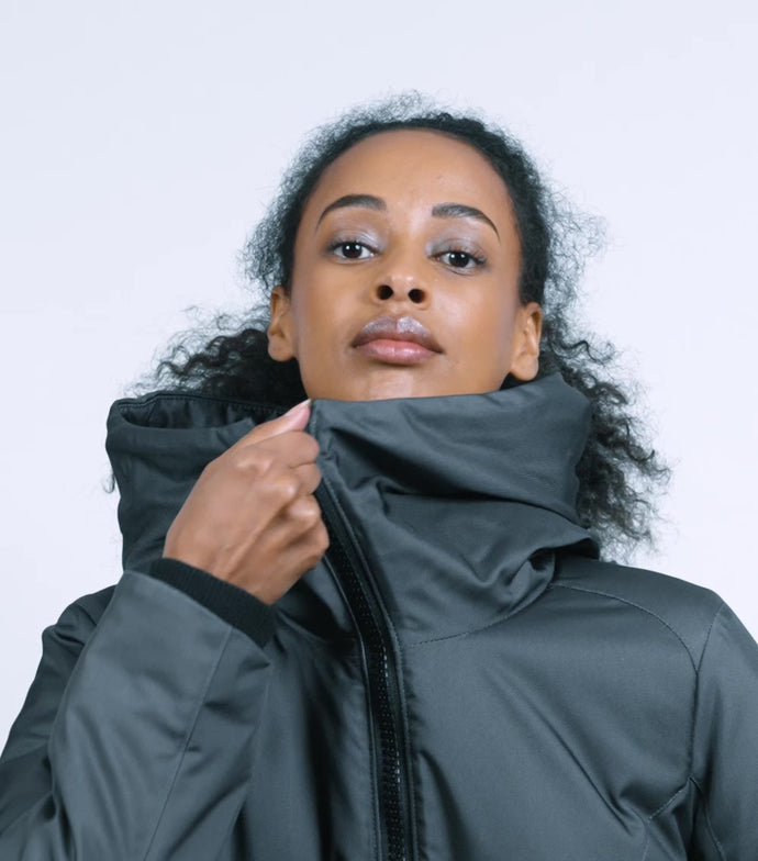 A woman zips up a Bedi coat, and then raises the hood over her head. She then turns sideways for a profile view