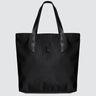A black nylon tote bag with black medium-length straps and a black patch with gold logo text