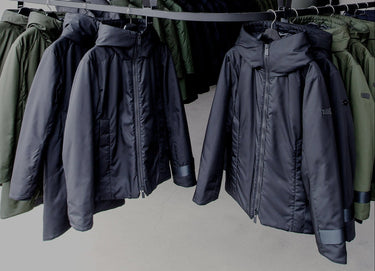 A dark navy hooded puffer jacket hanging on a black rack, flanked by olive green and black jackets on hangers in a store or warehouse setting.